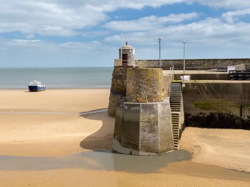 A picturesque view of the Saundersfoot Lighthouse standing tall against a clear blue sky, with gentle waves lapping against the sandy shoreline below. The lighthouse, a symbol of maritime history, offering a serene coastal landscape.
